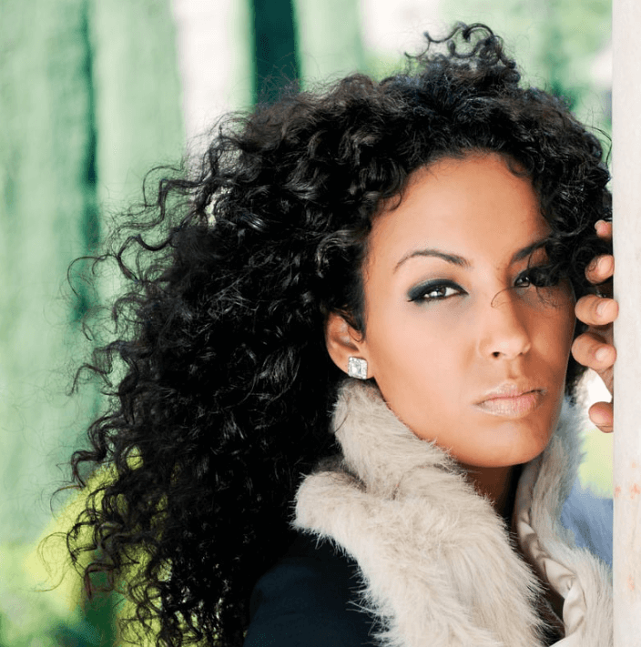 Mujer con cabello rizado posando, usando maquillaje de ojos, con un abrigo de piel, de fondo un entorno natural.